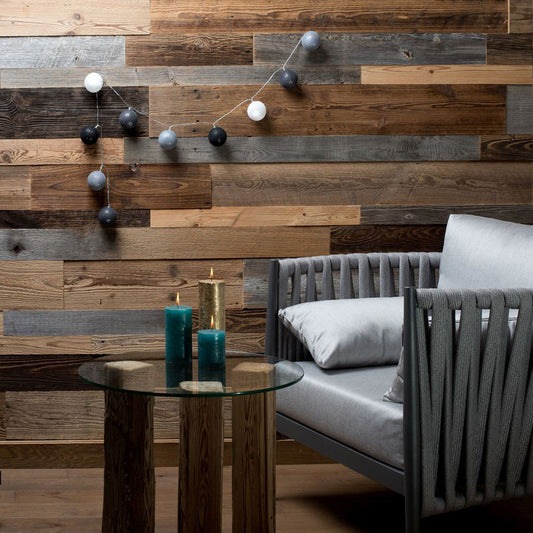 Une pièce moderne douillette mettant en vedette Wood & Wall Decor™ Parement Bois Antik, un fauteuil gris, une table ronde en verre, des bougies bleues et dorées, et des guirlandes lumineuses crée une atmosphère charmante. Produit présenté : Parement Bois Antik.