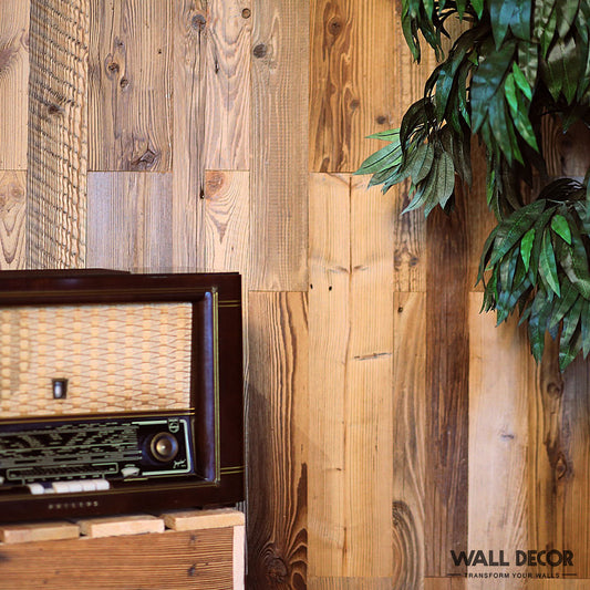 Le Parement Bois Chalet Alpine (0,5 m²) de Wood & Wall Decor™ est posé sur une surface en bois contre un mur rustique, accentué par des branches végétales feuillues. Cette décoration murale élégante apporte un charme nostalgique à n'importe quelle maison. "Décoration murale" apparaît en bas à droite.