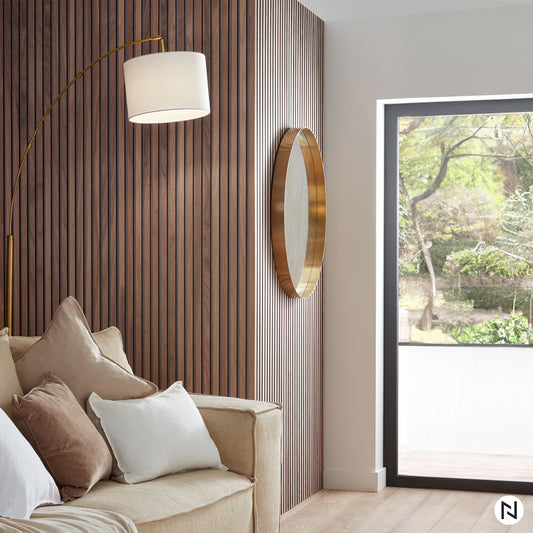 Un coin salon confortable avec un canapé beige, des coussins assortis, un lampadaire incurvé et un miroir rond encadré d'or présente le Panneau Tasseaux Bois Acoustiques - Noyer Naturel - Feutre gris de NatureWall™ pour le style et la sérénité au milieu de la vue sur le jardin.