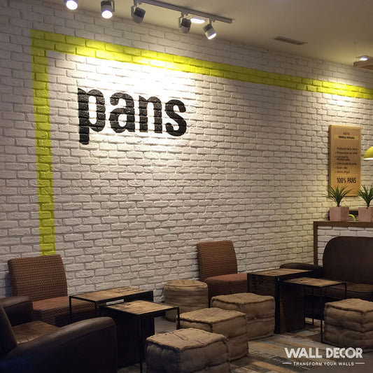 Un café moderne présentant Panespol™ Parement Brique blanche - Urban Brick sur les murs, avec des chaises brunes, des tables basses et des poufs. Le mot "pans" est peint en noir sur le mur de briques blanches, délimité par un liseré jaune.