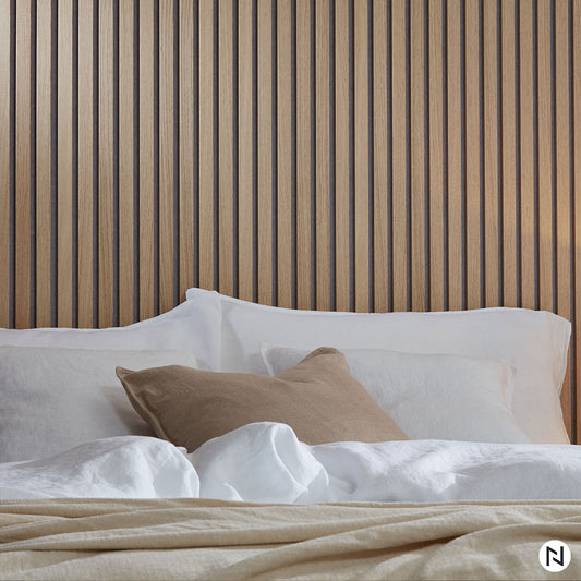 Un lit avec une literie blanche et beige repose sur un NatureWall™ Panneau Tasseaux Bois Acoustiques - Chêne Naturel - Feutre gris tête de lit, accentué par des oreillers blancs et un oreiller bronzé pour un style moderne et apaisant.