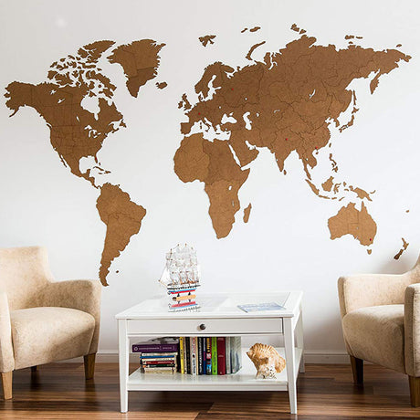 La Carte du monde en Bois 280x170cm - Marron - Géante by World Map™ est accrochée à un mur blanc au-dessus d'une table basse avec des livres, un coquillage et une maquette de bateau, flanquée de fauteuils beiges sur des planchers en bois-créant une harmonie équilibrée dans votre espace.