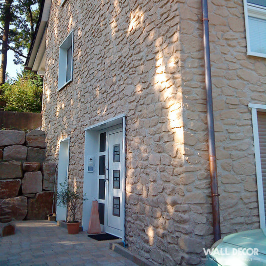 L'extérieur d'une maison présente Covering Panels™ Plaque Pierre Moellon Un revêtement en pierre beige (2,85 m x 1,3 m, 3,7 m²) de Pierres Ginestar, une porte blanche à panneaux de verre, deux fenêtres, une plante en pot près de l'entrée et un mur de soutènement en pierres empilées sur la gauche.