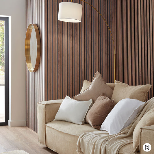 Un canapé beige confortable avec des coussins assortis est assis contre un mur d'accent NatureWall™ Panneau Tasseaux Bois Acoustiques - Noyer Naturel, sous un miroir rond encadré d'or et un lampadaire moderne arqué, avec la lumière naturelle qui coule à flot depuis la gauche.
