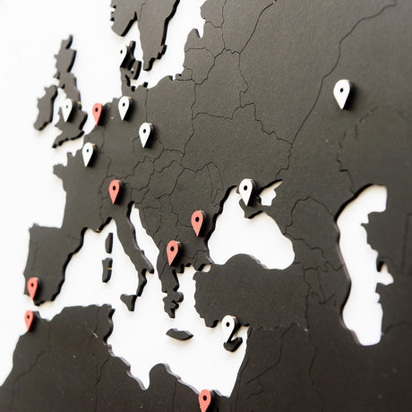 La World Map™ Carte du monde en Bois 180x108cm - Noir est une carte de l'Europe en bois noir et blanc, dotée d'un effet 3D unique avec des épingles rouges et blanches mettant en évidence différents pays et villes.