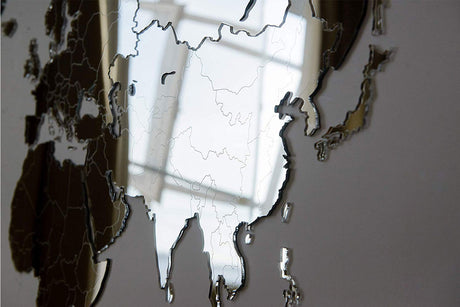 Un gros plan de la World Map™ Carte du monde en Bois 130x78cm - Miroir met en évidence le contour de l'Asie, reflétant la lumière naturelle et certaines parties de la pièce dans sa surface miroir.