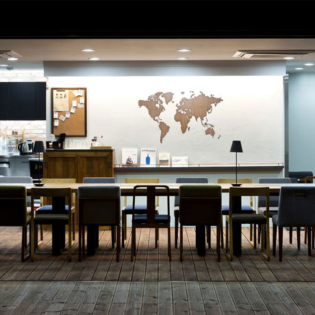Un intérieur de café moderne avec des tables et des chaises en bois, un comptoir avec des livres et une lampe, un tableau en liège sur le mur de gauche, et la World Map™ Carte du monde en Bois 130x78cm - Noyer - Exclusive comme art mural frappant - le tout dans un style minimaliste.