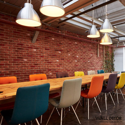 Une longue table en bois avec des chaises colorées est placée devant un mur Panespol™ Parement Brique rouge - Urban Brick, sous des lumières industrielles dans cet espace de travail moderne, où des meubles vibrants et de la verdure ressortent dans la lumière naturelle.