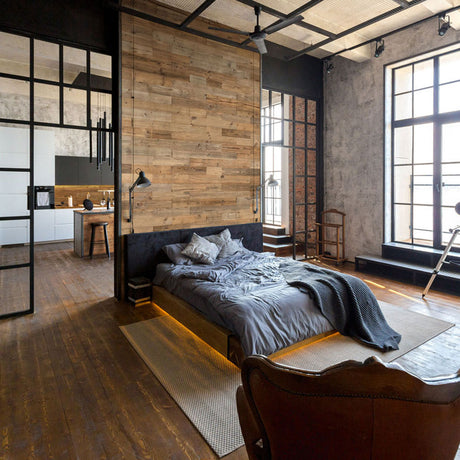 Une chambre à coucher moderne présente un mur d'accent Parement Bois Nature de Wood & Wall Decor™, de grandes fenêtres, une literie grise et un agencement ouvert menant à la cuisine. Un éclairage chaleureux sous le lit crée une atmosphère douillette de style industriel.