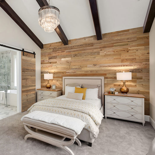 Une chambre à coucher confortable avec un mur d'accent Wood & Wall Decor™ Parement Bois Grange, un plafond voûté avec des poutres sombres, une literie neutre avec un oreiller jaune, deux tables de nuit avec des lampes, un banc et une porte de grange coulissante menant à la salle de bain.