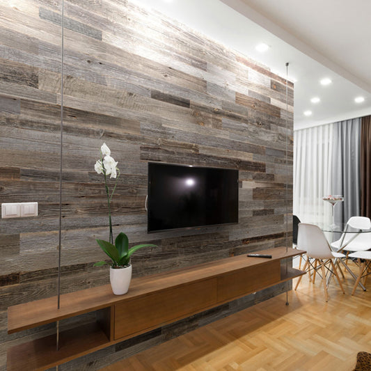 Salon moderne présentant un mur d'accent Wood & Wall Decor™ Parement Bois Anthracite, une télévision murale, une longue étagère en bois avec une orchidée blanche, et un coin repas avec des chaises blanches en arrière-plan.