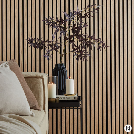 Un vase noir avec des branches feuillues sombres repose sur une petite étagère noire à côté de deux bougies blanches allumées et de livres. Derrière, NatureWall™ Panneau Tasseaux Bois Acoustiques - Chêne Naturel forme une toile de fond moderne, avec un canapé beige que l'on aperçoit en partie sur la gauche.