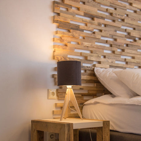 Le Parement Bois Visio de Wood & Wall Decor™ présente une lampe de chevet moderne avec un abat-jour noir sur une table de nuit en bois à côté d'oreillers blancs et d'une tête de lit en bois texturé, mêlant style et fonction pour votre chambre à coucher.