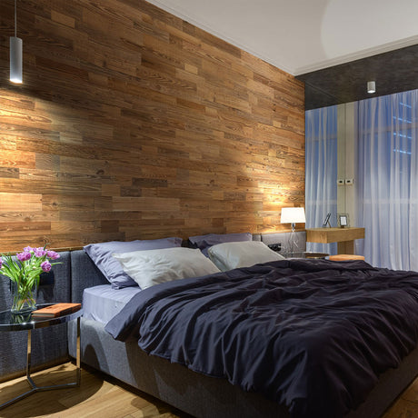 Chambre à coucher moderne avec un mur d'accent Parement Bois Grange Lite de Wood & Wall Decor™, un lit capitonné gris, une literie foncée, une table de nuit avec des fleurs et un livre, ainsi qu'un bureau et une chaise près de grandes fenêtres avec des rideaux diaphanes.