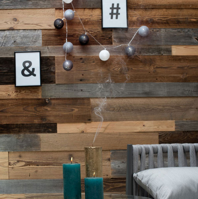 Un Parement Bois Antik de Wood & Wall Decor™ est posé sur une table avec deux bougies sarcelle et un vase or avec de la fumée devant une chaise coussinée grise, créant un décor élégant et moderne parfait pour les intérieurs contemporains.