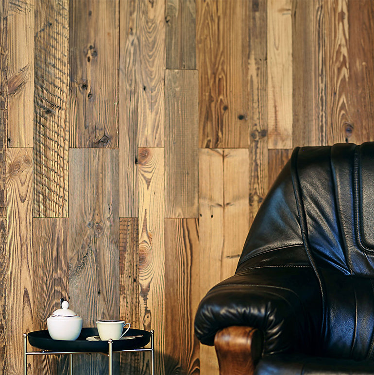 Un fauteuil de repos en cuir noir est posé à côté d'une petite table d'appoint ronde avec une théière et une tasse à thé blanches, posées contre un Wood & Wall Decor™ Parement Bois Chalet Alpine - 0,5 m² de mur en bois rustique.