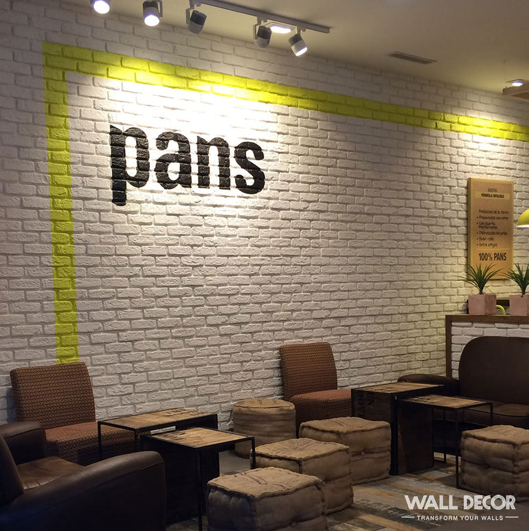 Un café moderne présentant Panespol™ Parement Brique blanche - Urban Brick sur les murs, avec des chaises brunes, des tables basses et des poufs. Le mot 