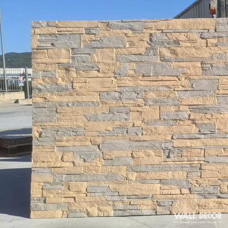 En extérieur, à la lumière du jour, la Plaque Pierre Alpine by Covering Panels™ présente des panneaux beige et gris effet pierre (2,85m x 1,3m : 3,7m²). Le sol pavé mène à un bâtiment industriel avec des montagnes au-delà. La mention "WALL DECOR" apparaît en bas à droite.
