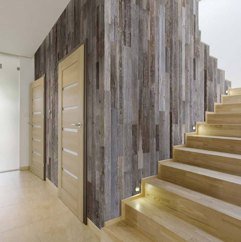 Un couloir moderne avec des escaliers en bois clair dotés de lampes de marche intégrées, un sol en carrelage beige, deux portes en bois clair et un mur orné de Parement Bois Anthracite de Wood & Wall Decor™ dans une disposition verticale rustique.