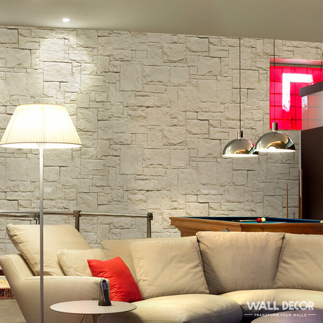 Un espace de vie confortable avec un canapé sectionnel beige, un coussin d'accent rouge, un lampadaire et un mur à texture de pierre claire fini en Covering Panels™ Plaque Pierre crème - Style Travertin (2,85m x 1,3m : 3,7 m²). Une table de billard et des lampes suspendues métalliques se trouvent en arrière-plan.