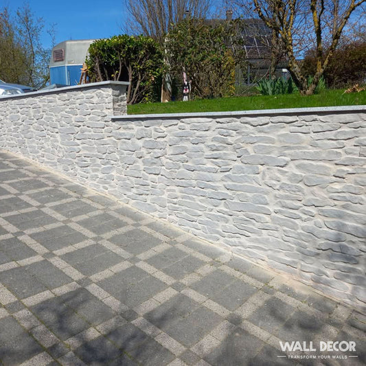 La Plaque Pierre grise - Pierres gneiss Ascona (2,85m x 1,3m : 3,7 m²) de Covering Panels™ forme un élégant mur de pierres grises à côté d'un dallage en damier, avec de l'herbe et des arbres au-delà sous la lumière du soleil et des ombres claires.