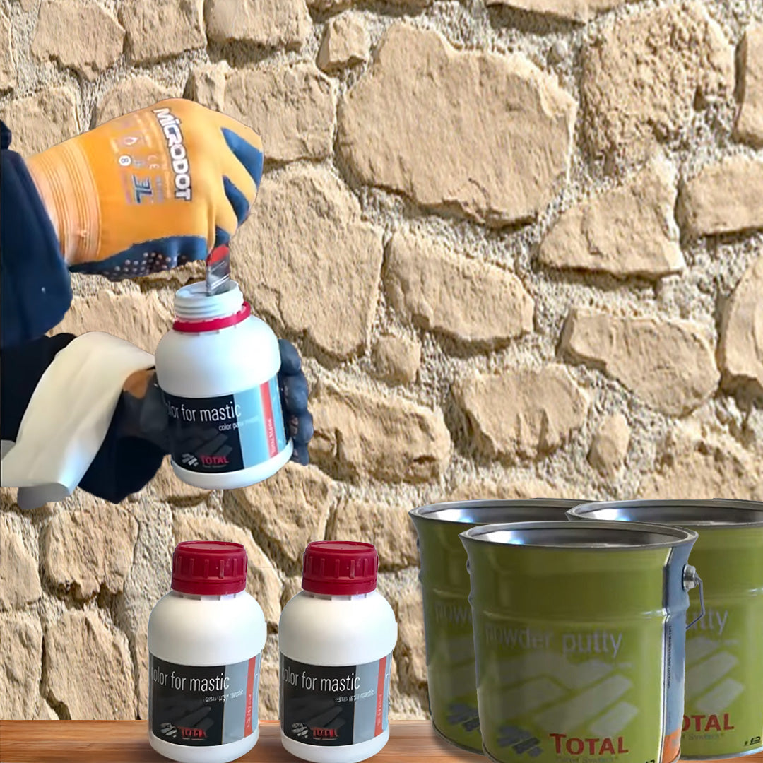 Une personne portant des gants orange ouvre un conteneur blanc étiqueté "Kit de Finition Parement Mural" par Covering Panels™ sur une surface grise, avec deux autres conteneurs et trois pots de peinture verts à proximité, posés contre un mur en pierre.
