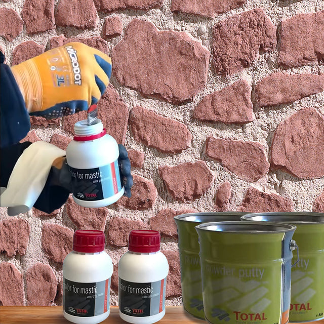 Une personne portant des gants de protection ouvre un petit récipient de Covering Panels™ Kit de Finition Parement Mural près d'un mur en pierre. Sur une table grise, il y a deux autres récipients identiques et trois bidons verts du kit.
