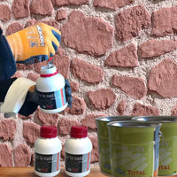 Une personne portant des gants de protection ouvre un petit récipient de Covering Panels™ Kit de Finition Parement Mural près d'un mur en pierre. Sur une table grise, il y a deux autres récipients identiques et trois bidons verts du kit.