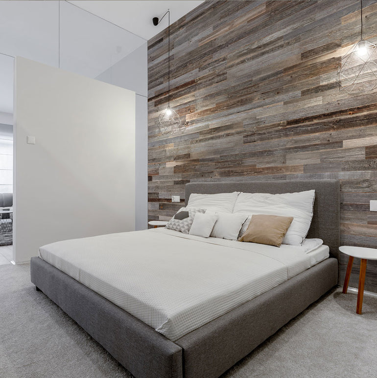 Chambre à coucher moderne avec un lit capitonné gris, une literie blanche et des oreillers neutres, avec un mur d'accent rustique Wood & Wall Decor™ Parement Bois Anthracite, deux lampes suspendues et des tables de nuit blanches de chaque côté.