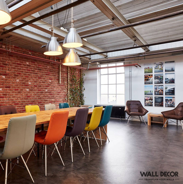 Salle de réunion moderne avec une longue table en bois, des chaises colorées, un plafond industriel, une grande fenêtre, de l'art mural et des lampes suspendues. Le mur en briques apparentes utilise Parement Brique rouge - Urban Brick de Panespol™, ce qui renforce l'atmosphère vibrante.