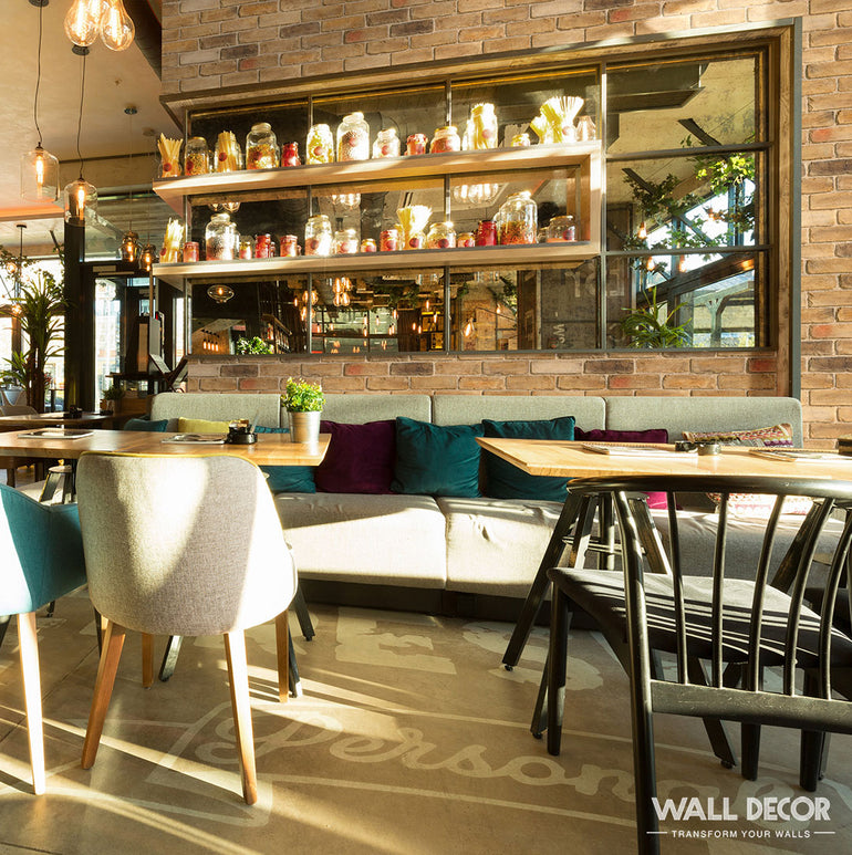 Un café confortable présente des murs en Panespol™ Parement Brique Marron - English Brick, un canapé rembourré, des coussins colorés, des tables en bois, des chaises mixtes et des étagères avec de la décoration. De grands miroirs reflètent la lumière du soleil - parfaits pour les séances de travail ou d'étude.