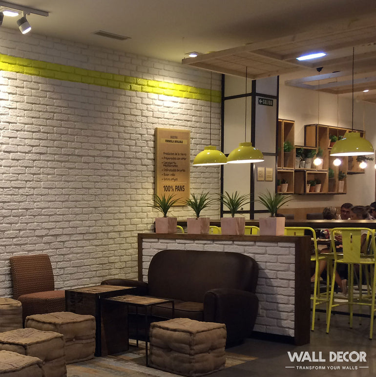 L'intérieur d'un café moderne présente des murs Panespol™ Parement Brique blanche - Urban Brick, des canapés bruns, des ottomans cubiques, des chaises jaunes et des plantes en pot sur une cloison sous des lampes suspendues.