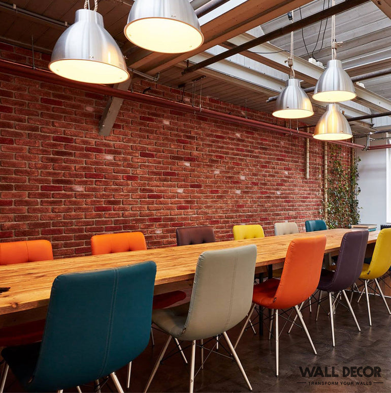 Une longue table en bois avec des chaises colorées est placée devant un mur Panespol™ Parement Brique rouge - Urban Brick, sous des lumières industrielles dans cet espace de travail moderne, où des meubles vibrants et de la verdure ressortent dans la lumière naturelle.