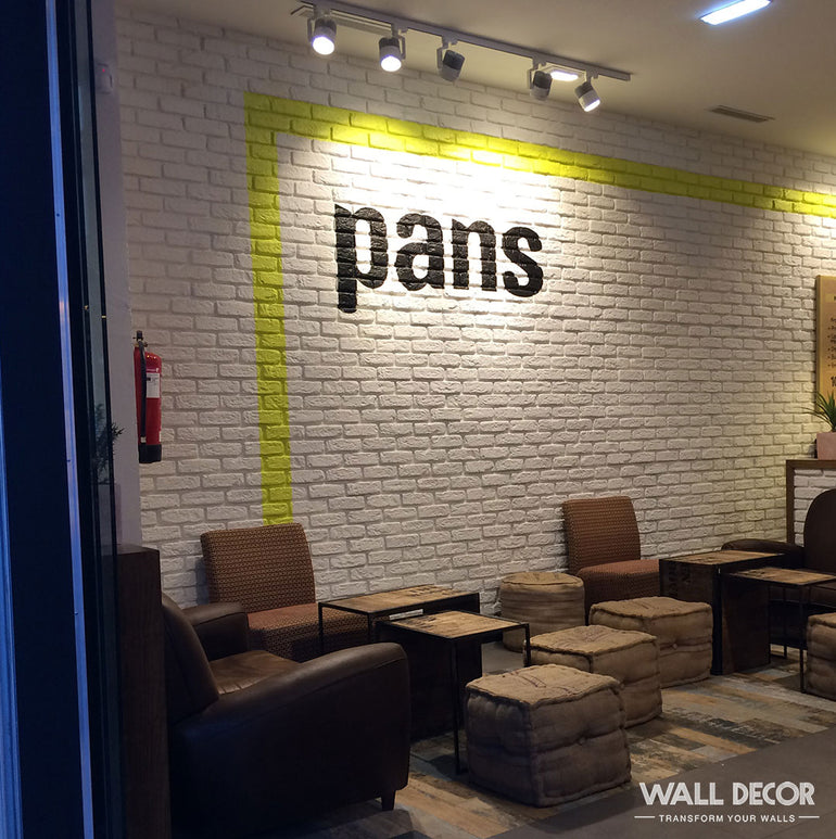 Un espace café confortable avec des chaises en osier et des tabourets coussinés s'appuie sur un mur Parement Brique blanche - Urban Brick de Panespol™, créant une ambiance accueillante et élégante, rehaussée par des planchers en bois et des détails de conception modernes.