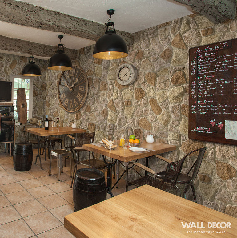 Un café confortable avec des murs en pierre Panespol™ Parement Pierre Meuliere - Cuenca, des tables en bois, des chaises noires, des lampes industrielles, une grande horloge, un menu sur tableau noir et des tables avec des assiettes, des verres, des fruits et une théière sur un sol carrelé.