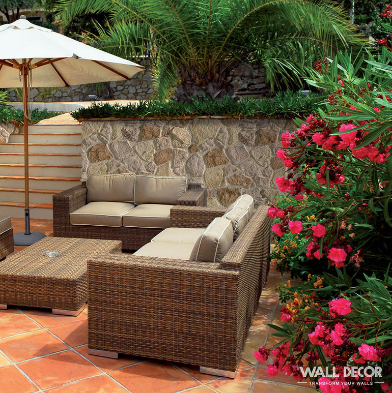 Des meubles de patio en osier avec des coussins beiges sont assis sur une terrasse carrelée sous un parasol. Des buissons à fleurs roses et des palmiers entourent l'espace, avec en arrière-plan d'élégants murs en pierre Panespol™ Parement Pierre Meuliere - Cuenca et des marches en pierre.