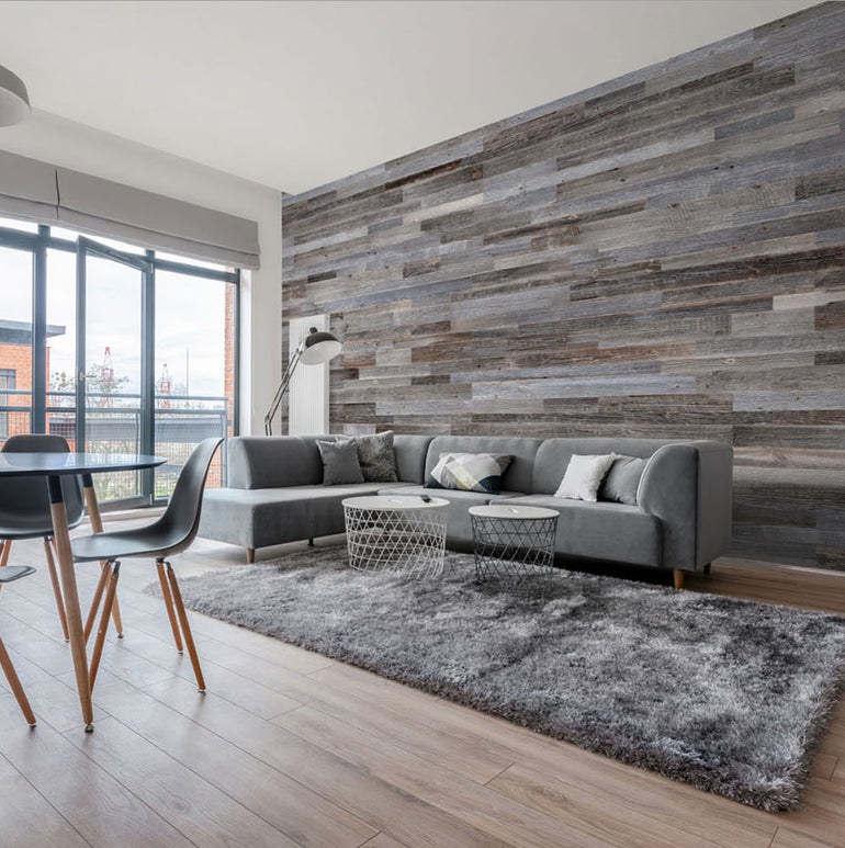 Un salon moderne présente un sectionnel gris, des tables basses rondes et un tapis shaggy face à un élégant mur d'accent Parement Bois Anthracite de Wood & Wall Decor™. De grandes fenêtres éclairent l'espace et révèlent un balcon extérieur ; des chaises de salle à manger se trouvent au premier plan.