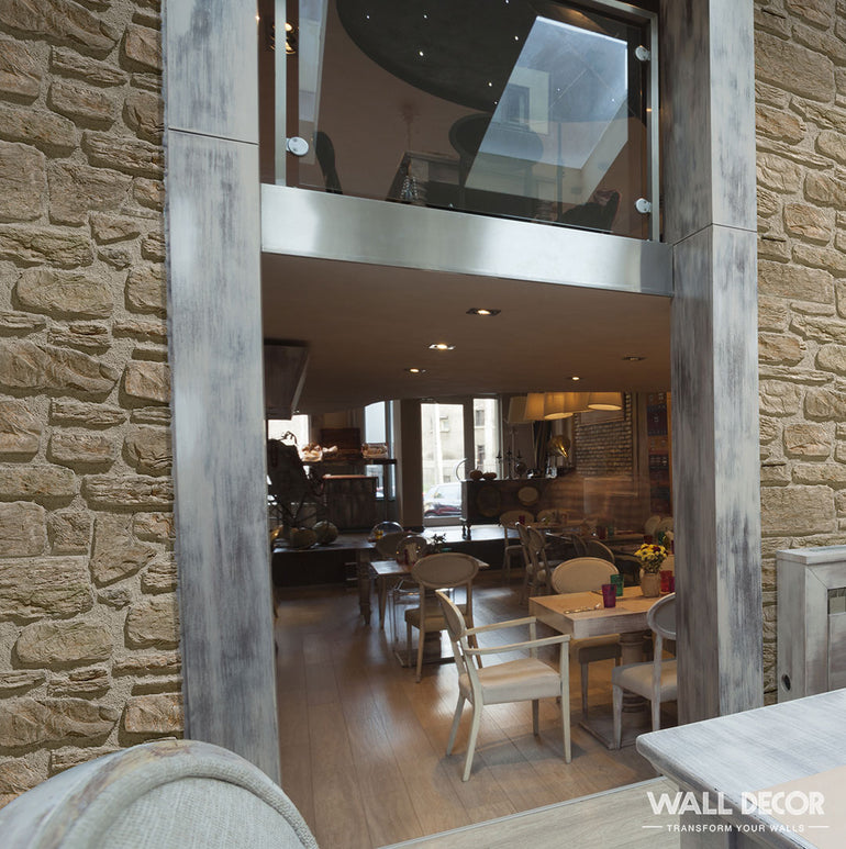 Un intérieur de café confortable présente Panespol™ Parement Pierre Galicia sur des murs en pierre rustique, des sols en bois et des meubles de style vintage, doucement éclairés par des plafonniers. Une mezzanine moderne en verre au-dessus ajoute de l'inspiration à la description du produit.