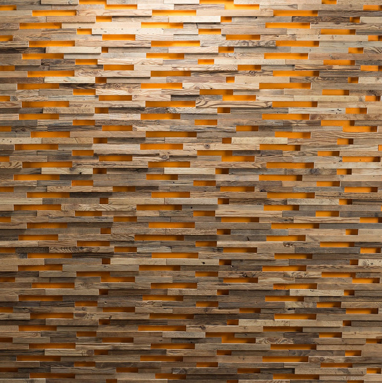 Parement Bois Visio by Wood & Wall Decor™ présente des planches de bois empilées de manière irrégulière avec des interstices, permettant à la lumière orange chaude de briller à travers et créant un aspect de mur texturé et stratifié.