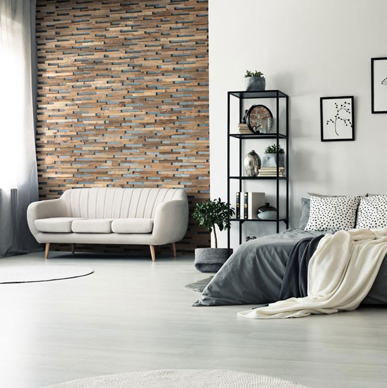 Un salon et une chambre modernes avec un canapé beige, une literie grise, des étagères en métal noir, des plantes et un mur d'accent fini en Parement Bois Visio par Wood & Wall Decor™. La lumière du soleil à travers la fenêtre apporte de la chaleur à l'espace minimaliste douillet.