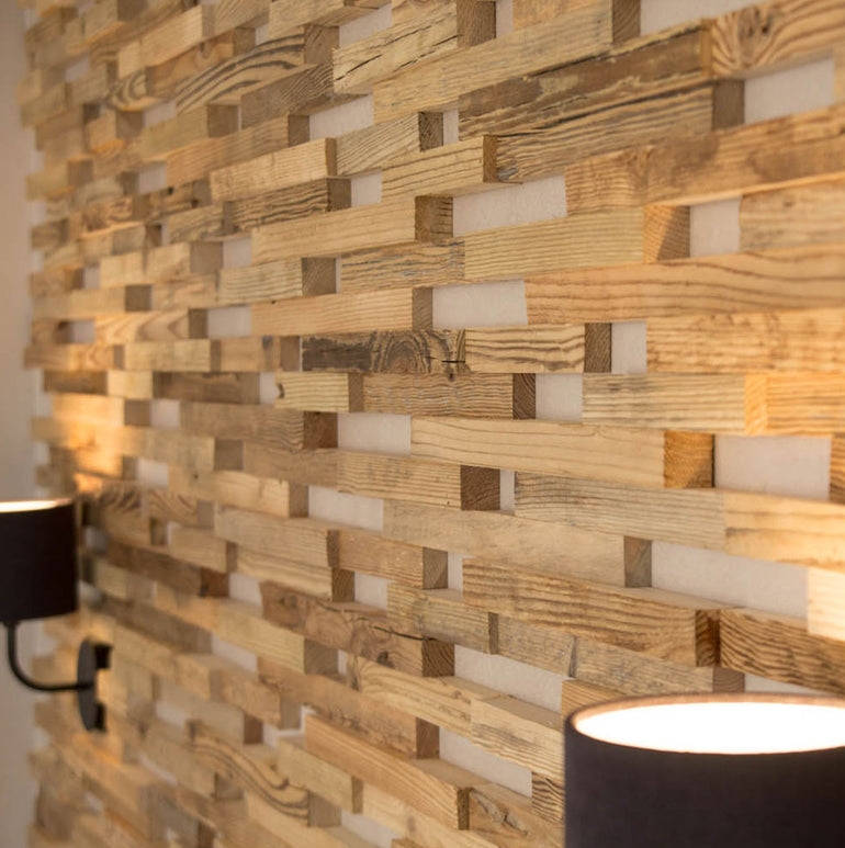 Un gros plan du Parement Bois Visio de Wood & Wall Decor™ montre son motif géométrique en bois en quinconce dans des tons naturels. Deux appliques murales aux nuances noires diffusent une lumière chaude, mais aucun code HTML ou CSS n'apparaît dans cette description de produit.