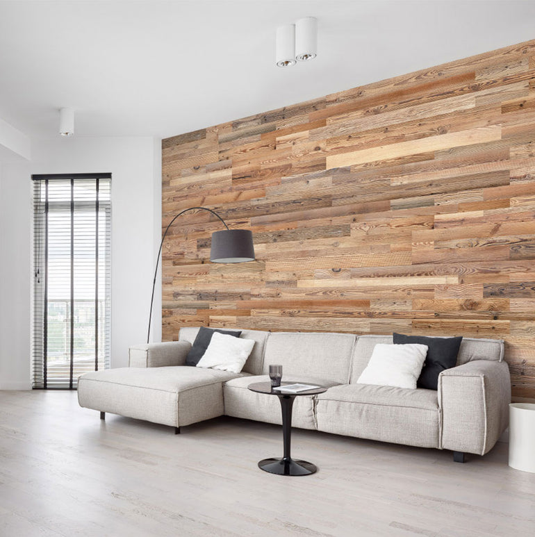 Un salon moderne présente un canapé sectionnel beige, une table basse ronde noire, un lampadaire haut et un mur d'accent Wood & Wall Decor™ Parement Bois Grange. De grandes fenêtres avec stores et des sols en bois clair renforcent l'ambiance lumineuse.