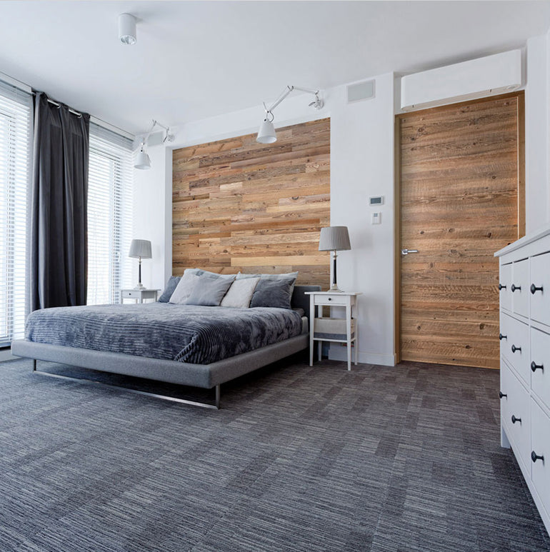 Chambre à coucher moderne avec un lit gris, plusieurs oreillers, deux tables de chevet assorties avec lampes, un mur et une porte d'accent Wood & Wall Decor™ Parement Bois Grange, une grande fenêtre avec stores, et une commode blanche sur une moquette grise.