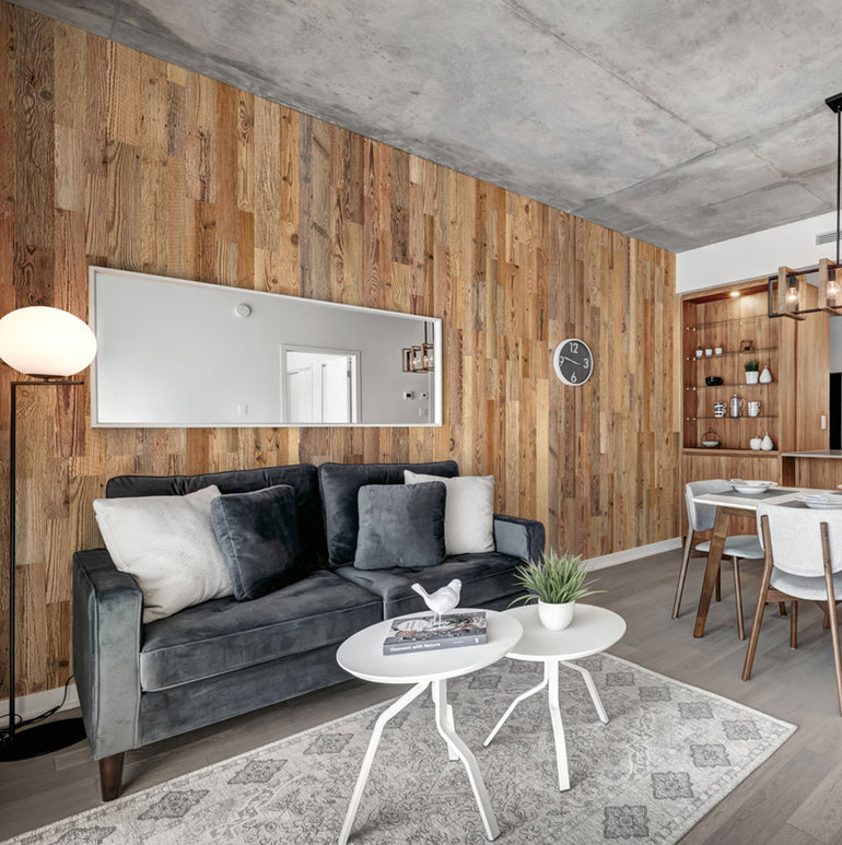Salon moderne composé d'un canapé gris, de coussins décoratifs, de tables basses rondes blanches, d'un tapis à motifs et d'un mur d'accent en panneaux de bois avec Parement Bois Grange de Wood & Wall Decor™. Un grand miroir, une horloge murale et un lampadaire complètent le look.