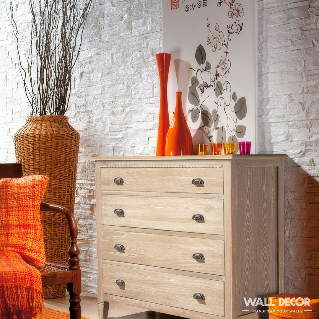 Une commode en bois clair avec des vases colorés à côté d'un jeté orange s'appuie sur un mur de briques blanches comportant Covering Panels™ Plaque Pierre Blanche - Laja (2,85m x 1,3m), ajoutant un style moderne à l'espace.