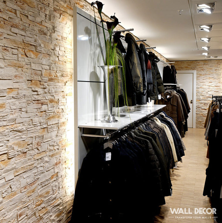 Un magasin de vêtements moderne présente des vestes et des manteaux sur des étagères contre un mur d'accent en pierre Panespol™ Parement Pierre Beige - Pizarra, avec un grand vase de plantes vertes présenté sur une étagère au-dessus.