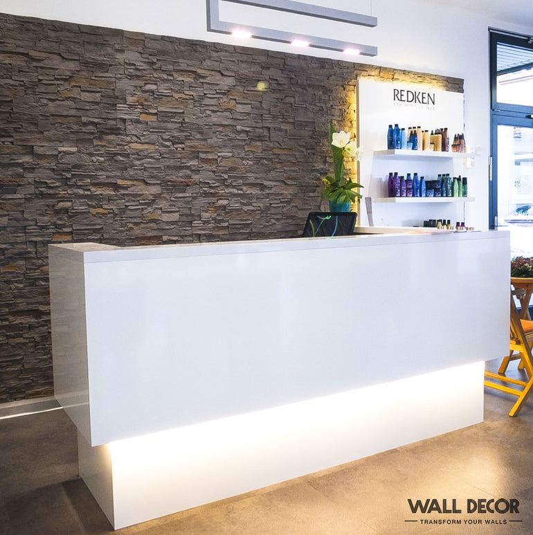 La réception d'un salon de coiffure moderne présente un bureau blanc épuré, des étagères contenant des produits capillaires et un mur d'accent en pierre fini en Panespol™ Parement Pierre Grise - Pizarra. L'espace minimaliste est égayé par un éclairage abondant et une plante en pot sur le comptoir.