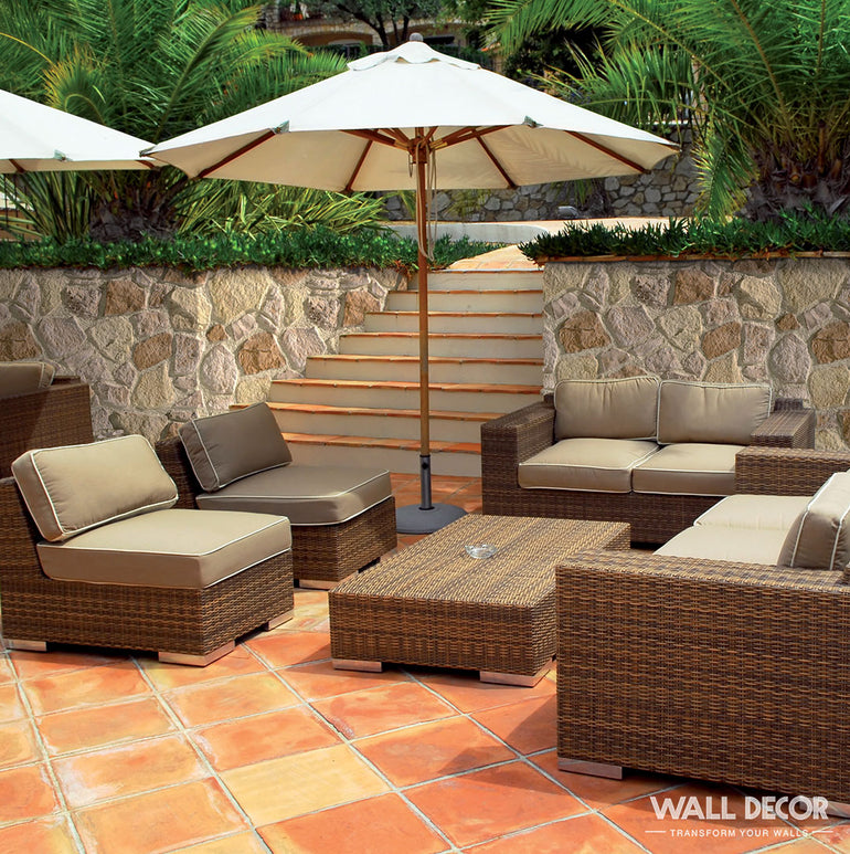 Le patio extérieur présente des panneaux muraux décoratifs Panespol™ Parement Pierre Meuliere - Cuenca, ajoutant du charme aux côtés de meubles en osier, de coussins beiges, d'une table basse et d'un grand parasol au milieu d'une végétation luxuriante.