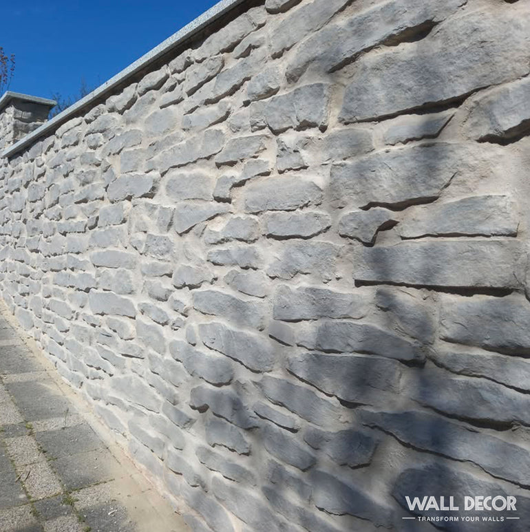 Un haut mur de pierres de gneiss gris clair (Plaque Pierre grise - Pierres gneiss Ascona, 2,85m × 1,3m : 3,7 m²) par Covering Panels™ longe un trottoir pavé sous un ciel bleu clair, avec une surface irrégulière et rustique.
