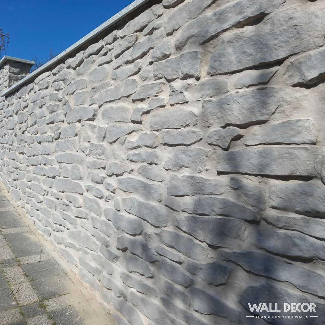 Un haut mur de pierres de gneiss gris clair (Plaque Pierre grise - Pierres gneiss Ascona, 2,85m × 1,3m : 3,7 m²) par Covering Panels™ longe un trottoir pavé sous un ciel bleu clair, avec une surface irrégulière et rustique.